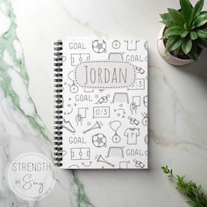 May include: A spiral-bound notebook with a white cover featuring a soccer-themed doodle pattern in black. The name "JORDAN" is displayed in a gray oval. The notebook is surrounded by a small potted plant and a marble surface.
