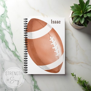 May include: A spiral-bound notebook with a watercolor illustration of a brown and white football. The name "Isaac" is printed in gray at the top. The notebook is on a white and marble surface. A small potted plant is in the background.