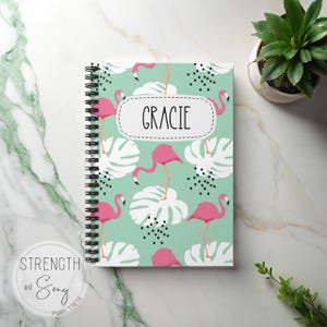 May include: A spiral-bound notebook with a flamingo pattern. The cover features pink flamingos, white leaves, and black dots on a light green background. The name "GRACIE" is displayed in a white oval. A small plant is in the background.