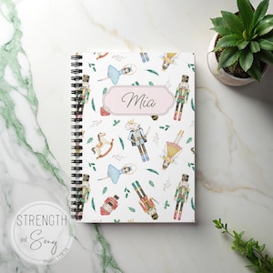 Puede incluir: Un cuaderno con espiral con una cubierta blanca con ilustraciones de acuarela de bailarinas, cascanueces y soldaditos de plomo. El nombre "Mia" está escrito en cursiva en una etiqueta rosa. El cuaderno está decorado con hojas verdes.