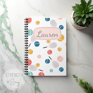 May include: A spiral-bound notebook with a white cover featuring a tennis theme. The cover is decorated with tennis balls and rackets in various colors, with the name "Lauren" in a pink oval. A small plant is in the background.