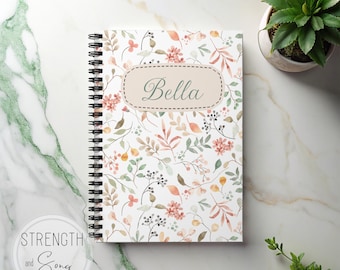 May include: A spiral-bound notebook with a floral pattern in shades of pink, orange, and green. The cover features the name "Bella" in an oval frame. The notebook is on a marble surface, with a small potted plant visible.