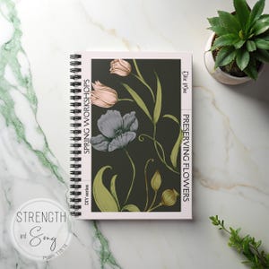 Op de afbeelding: Een spiraalgebonden notitieboek met een bloemenontwerp. De omslag heeft een donkere achtergrond met illustraties van roze en grijze bloemen en groene stelen. Tekst op de omslag luidt "Spring Workshops" en "Preserving Flowers". Een kleine potplant is zichtbaar.