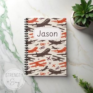 May include: Spiral-bound notebook with a cream background and a pattern of brown and orange airplanes. The name "Jason" is printed on a white label. The words "Strength and Song" and "Psalm 118:14" are printed on the bottom left.