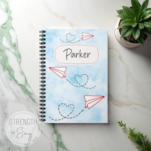 May include: A spiral-bound notebook with a light blue watercolor background and red paper airplane illustrations. The name "Parker" is written in a white oval. The notebook is on a marble surface with a small potted plant.