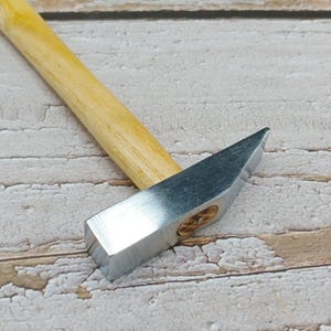 May include: A small, silver-colored hammer with a wooden handle. The hammer is lying on a white wooden surface.
