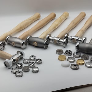 May include: A collection of metal hammers with wooden handles and various textured heads. Several interchangeable metal hammer heads with different patterns are also displayed, along with two white plastic hammer heads. The tools are for jewelry making.