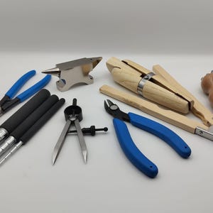 May include: A selection of jewellery-making tools, including pliers with blue handles, a small anvil, wooden clamps, and a compass. The tools are arranged on a white surface, showcasing the essential equipment for jewellery design and repair.