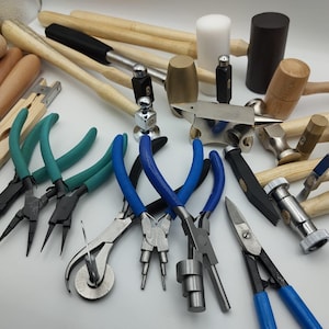 Könnte beinhalten: Eine Sammlung von Werkzeugen zur Schmuckherstellung, darunter Zangen mit blauen und grünen Griffen, Hämmer, Schlägel und ein kleiner Amboss. Verschiedene Werkzeuge haben Holzgriffe. Die Werkzeuge sind auf einer weißen Oberfläche angeordnet.