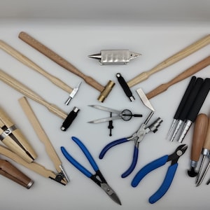 Könnte beinhalten: Eine Sammlung von Werkzeugen zur Schmuckherstellung, darunter Hämmer, Zangen und Dorne, angeordnet auf einer weißen Oberfläche. Die Werkzeuge haben Holzgriffe, silberne Metallköpfe und blaue Griffe. Ein Amboss ist ebenfalls vorhanden.