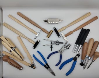 May include: A selection of jewellery-making tools, including hammers, pliers, and mandrels, arranged on a white surface. The tools feature wooden handles, silver metal heads, and blue handles. An anvil is also present.