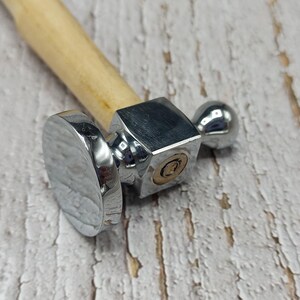 Op de afbeelding: Close-up van een metalen hamer met een houten handvat. De hamer heeft een gepolijste, zilverkleurige kop met een plat, rond slagvlak aan één kant, een vierkant midden en een afgerond uiteinde.