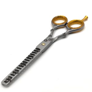May include: Silver and gold-toned pet grooming shears with paw print accents. The shears have a serrated edge and gold-coloured finger holes. The design is for pet grooming, with a focus on detail and functionality.