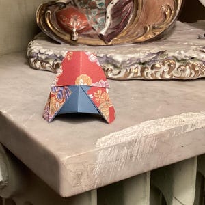 Peut inclure: Un petit objet en papier plié, rouge, bleu et motifs floraux dorés, est posé sur une surface en marbre. Une figurine décorative en porcelaine de deux personnes en costume du XVIIIe siècle est en arrière-plan.