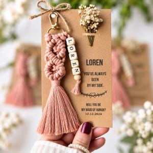 Op de afbeelding: Een oudroze macramé sleutelhanger met een goudkleurige karabijnhaak en houten kralen met de letters LOREN. De sleutelhanger is bevestigd aan een bruine kaart met de tekst LOREN YOU'VE ALWAYS BEEN BY MY SIDE. WILL YOU BE MY MAID OF HONOR? 12.12.2024 en een klein takje gedroogde bloemen.