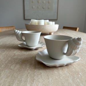 Peut inclure: Deux tasses à thé et soucoupes en céramique blanche avec des anses en forme de coquillage. Les tasses sont posées sur une surface claire et texturée. Un bol en bois avec des bougies blanches et une œuvre encadrée sont en arrière-plan.