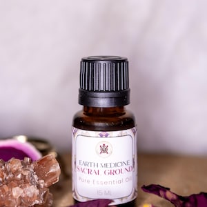 May include: A brown glass bottle with a black cap, labeled "Earth Medicine Sacral Ground Pure Essential Oil 15 ML." The label has a white background with purple text and a decorative border. Dried rose petals and crystals are in the background.