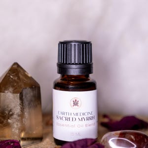 May include: A brown glass bottle with a black cap, labeled "EARTH MEDICINE SACRED MYRRH Essential Oil Blend." The bottle is 15 mL. A clear crystal and a polished stone are next to the bottle, with dried rose petals scattered around.