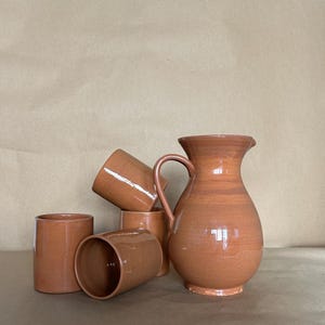 Puede incluir: Un juego de cuatro tazas de terracota y una jarra de terracota. Las tazas y la jarra son todas del mismo color, un marrón rojizo cálido.