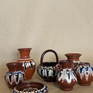 Puede incluir: Colección de cerámica de terracota hecha a mano. El conjunto incluye jarrones, una pequeña cesta con asa y un plato hondo. Cada pieza presenta una base marrón con diseños decorativos blancos, negros y azules. Medidas en centímetros.
