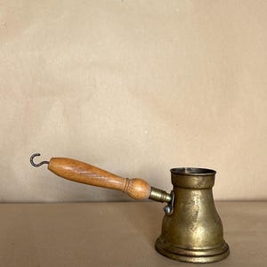 Peut inclure: Une cafetière en cuivre avec une poignée en bois. La cafetière a un corps arrondi et un bec verseur sur le côté. La poignée est fixée à la cafetière par un crochet en métal.