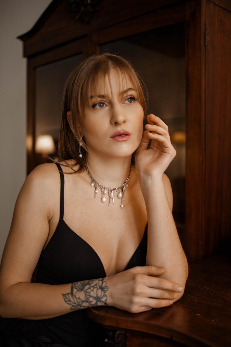 May include: A silver-toned chain necklace featuring multiple dangling white baroque pearls and small metallic charms. The necklace rests at the base of the neck and is paired with matching drop earrings featuring a single white pearl on a thin chain.