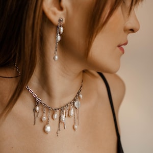 May include: A silver-toned chain necklace featuring multiple dangling white freshwater pearls of varying shapes and sizes, paired with a matching drop earring consisting of a silver chain and small white pearls hanging from a stud post.