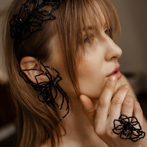 May include: A woman is wearing black beaded jewelry, including a headband, ear cuff, and ring. The jewelry has a floral design. The woman has light brown hair and is wearing a black top. The background is blurred.