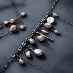 May include: A silver-toned metal chain necklace featuring multiple dangling charms, including various irregular-shaped cream-colored pearls and a single smooth, cylindrical metal bar. The design incorporates layered fine chains and larger links.