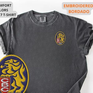 Mockup of a Comfort Colors 1717 vintage-colored t-shirt featuring a small embroidered Venezuela-inspired lion crest on the left chest. The embroidery showcases a gold lion inside a navy blue oval with red and white shield details and the year 1842. A close-up view of the embroidered design is displayed alongside the shirt, highlighting the textured stitching and premium craftsmanship. Ideal Venezuelan pride apparel, Venezuela gift, VZLA shirt, and Caracas-inspired clothing.