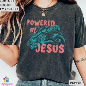 May include: Dark gray t-shirt with the text "POWERED BY JESUS" in pink, and a turquoise turtle graphic. The shirt also has the text "COMFORT COLORS 1717 T-SHIRT" in the upper left corner and the word "PEPPER" in the lower right corner.