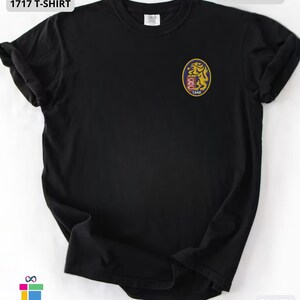Mockup of a Comfort Colors 1717 vintage-colored t-shirt featuring a small embroidered Venezuela-inspired lion crest on the left chest. The embroidery showcases a gold lion inside a navy blue oval with red and white shield details and the year 1842. A close-up view of the embroidered design is displayed alongside the shirt, highlighting the textured stitching and premium craftsmanship. Ideal Venezuelan pride apparel, Venezuela gift, VZLA shirt, and Caracas-inspired clothing.