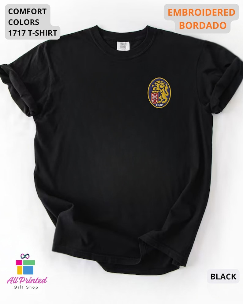 Mockup of a Comfort Colors 1717 vintage-colored t-shirt featuring a small embroidered Venezuela-inspired lion crest on the left chest. The embroidery showcases a gold lion inside a navy blue oval with red and white shield details and the year 1842. A close-up view of the embroidered design is displayed alongside the shirt, highlighting the textured stitching and premium craftsmanship. Ideal Venezuelan pride apparel, Venezuela gift, VZLA shirt, and Caracas-inspired clothing.