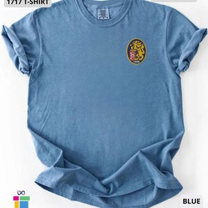 Mockup of a Comfort Colors 1717 vintage-colored t-shirt featuring a small embroidered Venezuela-inspired lion crest on the left chest. The embroidery showcases a gold lion inside a navy blue oval with red and white shield details and the year 1842. A close-up view of the embroidered design is displayed alongside the shirt, highlighting the textured stitching and premium craftsmanship. Ideal Venezuelan pride apparel, Venezuela gift, VZLA shirt, and Caracas-inspired clothing.