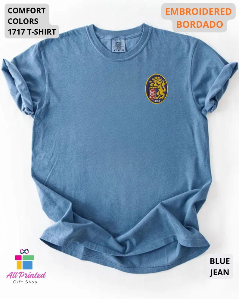 Mockup of a Comfort Colors 1717 vintage-colored t-shirt featuring a small embroidered Venezuela-inspired lion crest on the left chest. The embroidery showcases a gold lion inside a navy blue oval with red and white shield details and the year 1842. A close-up view of the embroidered design is displayed alongside the shirt, highlighting the textured stitching and premium craftsmanship. Ideal Venezuelan pride apparel, Venezuela gift, VZLA shirt, and Caracas-inspired clothing.
