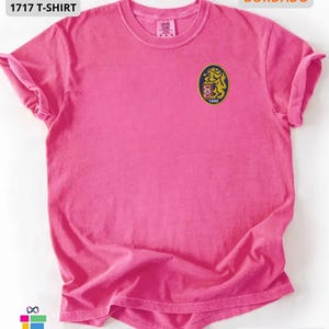 Mockup of a Comfort Colors 1717 vintage-colored t-shirt featuring a small embroidered Venezuela-inspired lion crest on the left chest. The embroidery showcases a gold lion inside a navy blue oval with red and white shield details and the year 1842. A close-up view of the embroidered design is displayed alongside the shirt, highlighting the textured stitching and premium craftsmanship. Ideal Venezuelan pride apparel, Venezuela gift, VZLA shirt, and Caracas-inspired clothing.