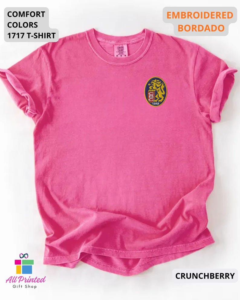 Mockup of a Comfort Colors 1717 vintage-colored t-shirt featuring a small embroidered Venezuela-inspired lion crest on the left chest. The embroidery showcases a gold lion inside a navy blue oval with red and white shield details and the year 1842. A close-up view of the embroidered design is displayed alongside the shirt, highlighting the textured stitching and premium craftsmanship. Ideal Venezuelan pride apparel, Venezuela gift, VZLA shirt, and Caracas-inspired clothing.