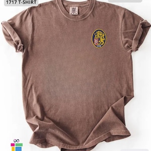 Mockup of a Comfort Colors 1717 vintage-colored t-shirt featuring a small embroidered Venezuela-inspired lion crest on the left chest. The embroidery showcases a gold lion inside a navy blue oval with red and white shield details and the year 1842. A close-up view of the embroidered design is displayed alongside the shirt, highlighting the textured stitching and premium craftsmanship. Ideal Venezuelan pride apparel, Venezuela gift, VZLA shirt, and Caracas-inspired clothing.