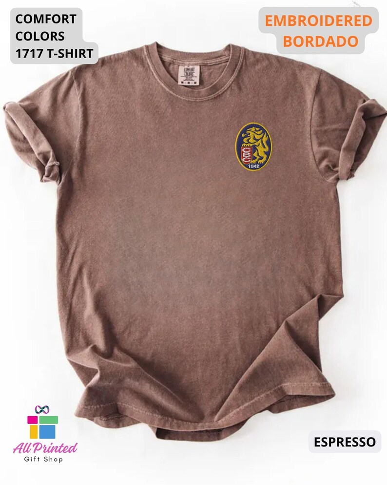 Mockup of a Comfort Colors 1717 vintage-colored t-shirt featuring a small embroidered Venezuela-inspired lion crest on the left chest. The embroidery showcases a gold lion inside a navy blue oval with red and white shield details and the year 1842. A close-up view of the embroidered design is displayed alongside the shirt, highlighting the textured stitching and premium craftsmanship. Ideal Venezuelan pride apparel, Venezuela gift, VZLA shirt, and Caracas-inspired clothing.