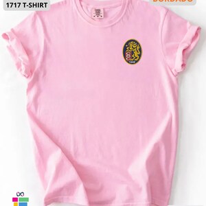 Mockup of a Comfort Colors 1717 vintage-colored t-shirt featuring a small embroidered Venezuela-inspired lion crest on the left chest. The embroidery showcases a gold lion inside a navy blue oval with red and white shield details and the year 1842. A close-up view of the embroidered design is displayed alongside the shirt, highlighting the textured stitching and premium craftsmanship. Ideal Venezuelan pride apparel, Venezuela gift, VZLA shirt, and Caracas-inspired clothing.