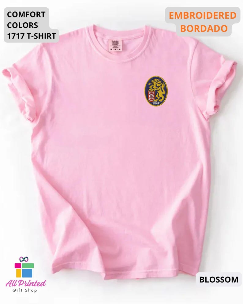 Mockup of a Comfort Colors 1717 vintage-colored t-shirt featuring a small embroidered Venezuela-inspired lion crest on the left chest. The embroidery showcases a gold lion inside a navy blue oval with red and white shield details and the year 1842. A close-up view of the embroidered design is displayed alongside the shirt, highlighting the textured stitching and premium craftsmanship. Ideal Venezuelan pride apparel, Venezuela gift, VZLA shirt, and Caracas-inspired clothing.