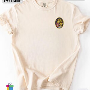 Mockup of a Comfort Colors 1717 vintage-colored t-shirt featuring a small embroidered Venezuela-inspired lion crest on the left chest. The embroidery showcases a gold lion inside a navy blue oval with red and white shield details and the year 1842. A close-up view of the embroidered design is displayed alongside the shirt, highlighting the textured stitching and premium craftsmanship. Ideal Venezuelan pride apparel, Venezuela gift, VZLA shirt, and Caracas-inspired clothing.
