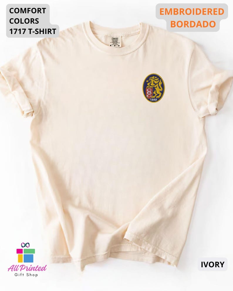 Mockup of a Comfort Colors 1717 vintage-colored t-shirt featuring a small embroidered Venezuela-inspired lion crest on the left chest. The embroidery showcases a gold lion inside a navy blue oval with red and white shield details and the year 1842. A close-up view of the embroidered design is displayed alongside the shirt, highlighting the textured stitching and premium craftsmanship. Ideal Venezuelan pride apparel, Venezuela gift, VZLA shirt, and Caracas-inspired clothing.