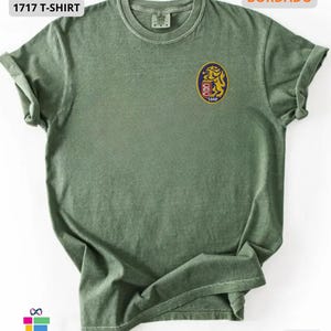 Mockup of a Comfort Colors 1717 vintage-colored t-shirt featuring a small embroidered Venezuela-inspired lion crest on the left chest. The embroidery showcases a gold lion inside a navy blue oval with red and white shield details and the year 1842. A close-up view of the embroidered design is displayed alongside the shirt, highlighting the textured stitching and premium craftsmanship. Ideal Venezuelan pride apparel, Venezuela gift, VZLA shirt, and Caracas-inspired clothing.