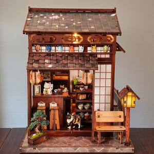 May include: A detailed wooden miniature Japanese house with a tiled roof, paper lanterns, and a small bench. The interior features miniature furniture, a bonsai tree, and a lucky cat figurine. Decorative masks are displayed above the entrance.