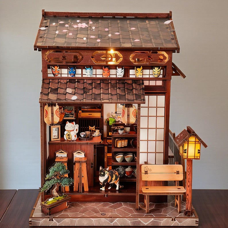 May include: A detailed wooden miniature Japanese house with a tiled roof, paper lanterns, and a small bench. The interior features miniature furniture, a bonsai tree, and a lucky cat figurine. Decorative masks are displayed above the entrance.