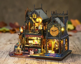 May include: A miniature multi-story building model featuring dark gray laser-cut walls, intricate gothic architectural details, and warm interior lighting. The structure includes a clock tower, a small fountain, and signage reading "be a real adventurer in the magical world."
