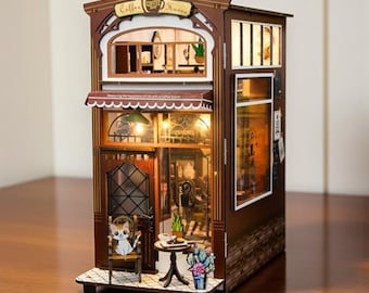 May include: A miniature wooden Coffee House model with intricate details. The two-story structure features a cafe with a small table and chair, a cat figurine, and a balcony. The sign reads "Coffee House". The model is illuminated with warm lighting.
