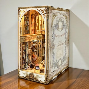 May include: A miniature, open-faced diorama of a library scene. The interior features bookshelves, books, and a reading area. The exterior is designed to look like a book with the title "Baroque Dreams" and decorative gold trim.