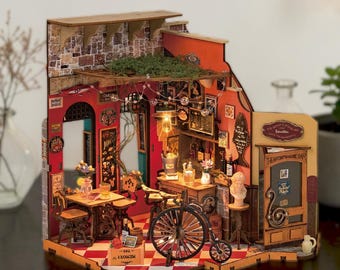 May include: A miniature wooden model of a bar interior featuring a red and white checkered floor, a vintage-style bicycle, a wooden table with chairs, a bar counter with bottles, a stone-textured wall, and a door labeled The Artist's House Bar.